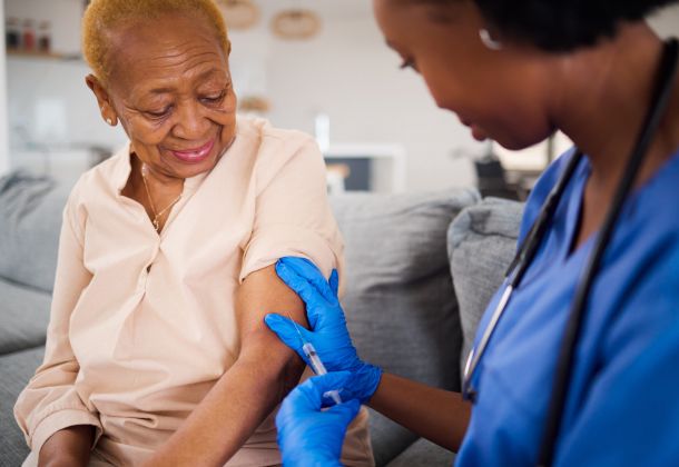 Smiling elderly woman receives a vaccine shot in her arm from a nurse wearing blue gloves.