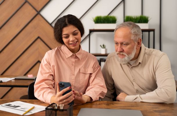 A young woman shows something on her phone to an older man as they sit together at a desk in a modern office.