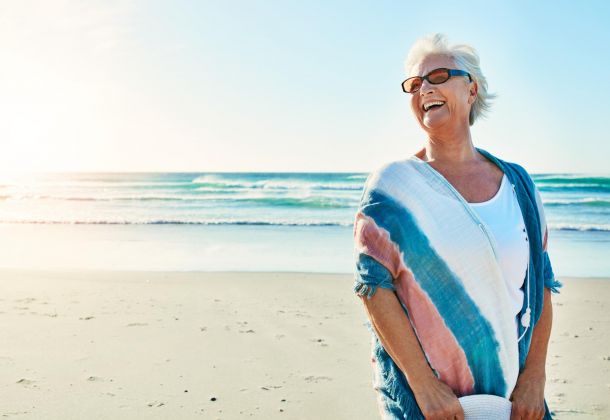 Smiling older woman in sunglasses stands on a sunny beach with waves in the background.