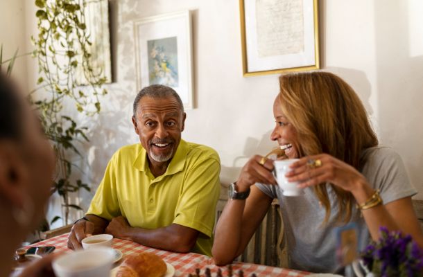 Two people sit at a table, smiling and drinking coffee together in a warmly lit room with framed art on the wall.