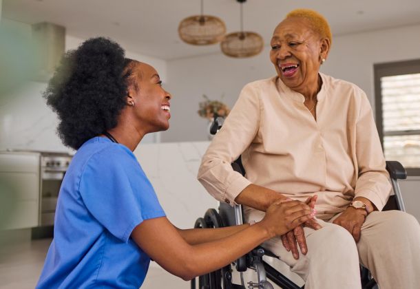 Nurse holding hands and smiling with an older woman in a wheelchair inside a bright home.