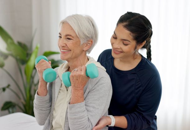 Elderly woman lifts dumbbells with assistance from a caregiver in a bright room.