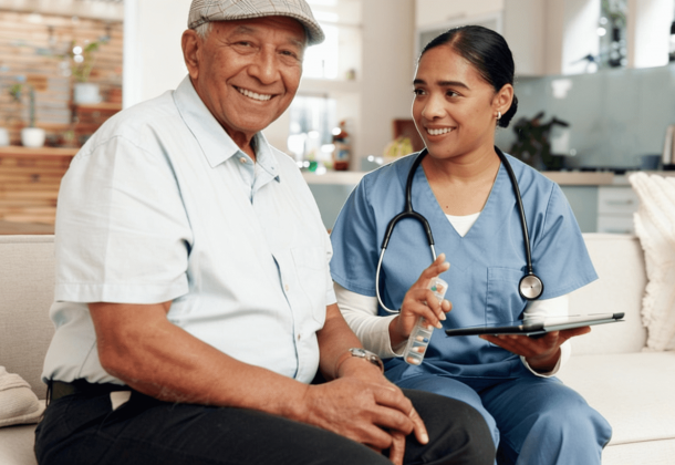 An elderly man smiles while sitting next to a nurse with a stethoscope and tablet in a bright living room.