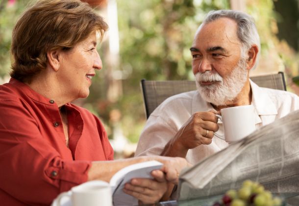 Smiling older couple sits outdoors, drinking coffee and reading a book and newspaper together.