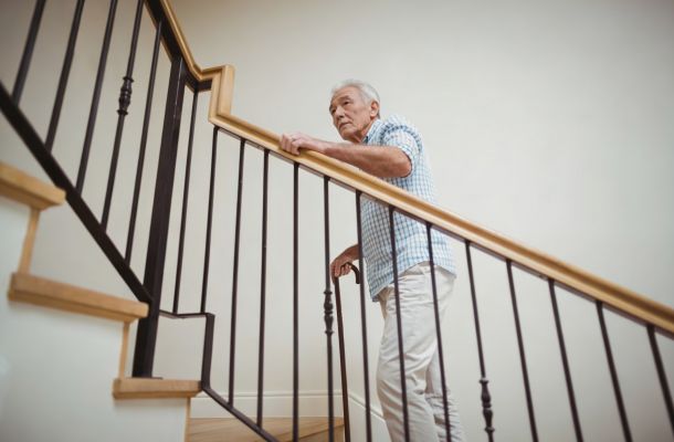 Older man in a checkered shirt holding a railing while walking up a staircase indoors.