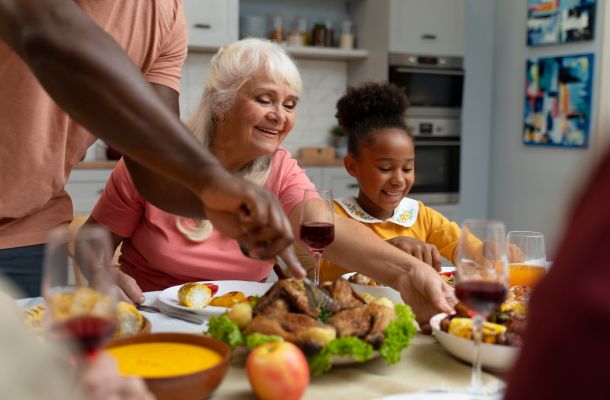 Smiling family sharing a meal at a table, with roast chicken, wine, and various dishes in a cozy kitchen.