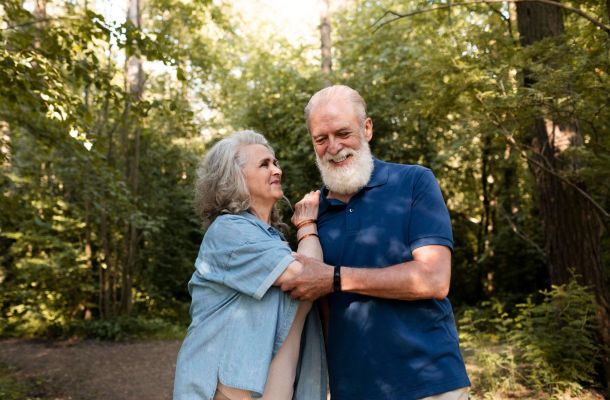 Smiling older couple standing together in a sunlit forest, enjoying each other's company.