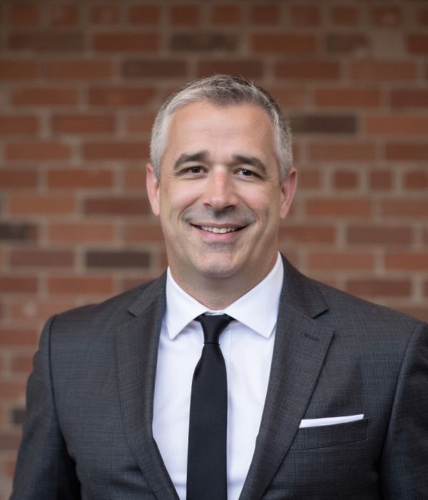 Smiling man in a suit and tie standing in front of a brick wall.