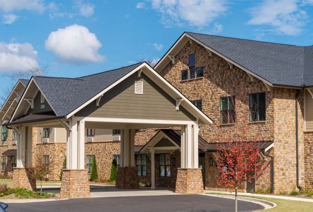 Brickmont at Johns Creek with stone facade, dark roof, covered entrance, and a small tree in front. Blue sky with clouds.