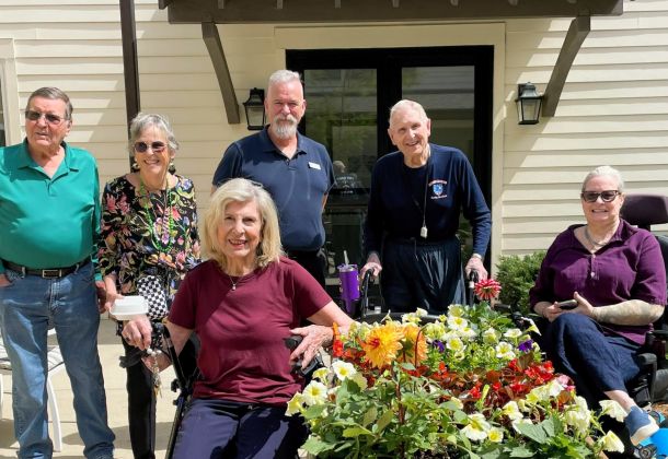 Six smiling older adults, some seated and some standing, pose outside by a colorful flower bed on a sunny day.
