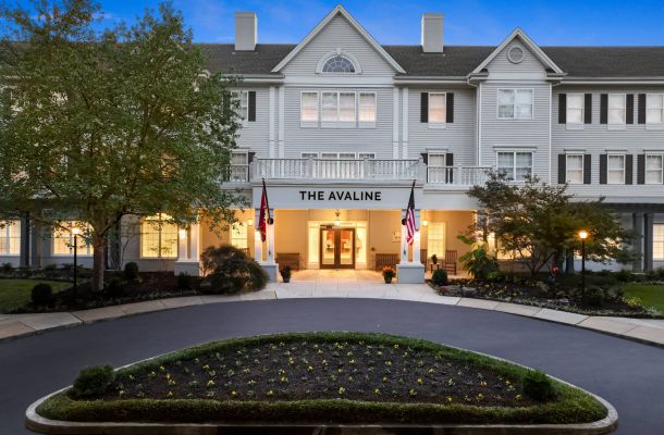 A large white senior living community named The Avaline with lit windows, two flags, and a landscaped driveway at dusk.