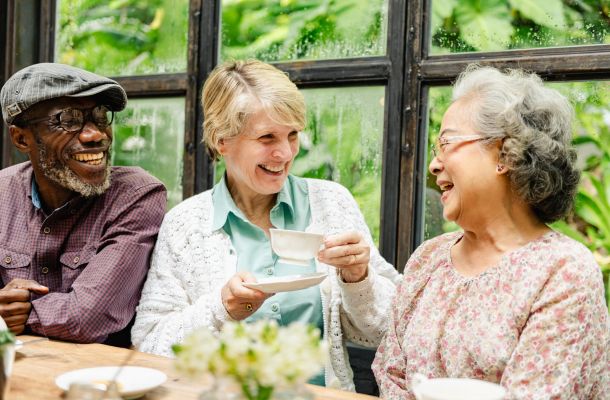 Three older adults laughing and enjoying tea together by a window with greenery outside.