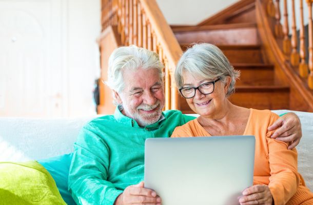 Smiling older couple sitting on a couch, looking at a laptop together in a cozy, sunlit living room with wooden stairs in the background.