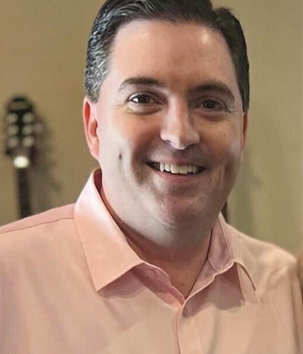 A smiling man in a light pink shirt stands indoors with guitars blurred in the background.