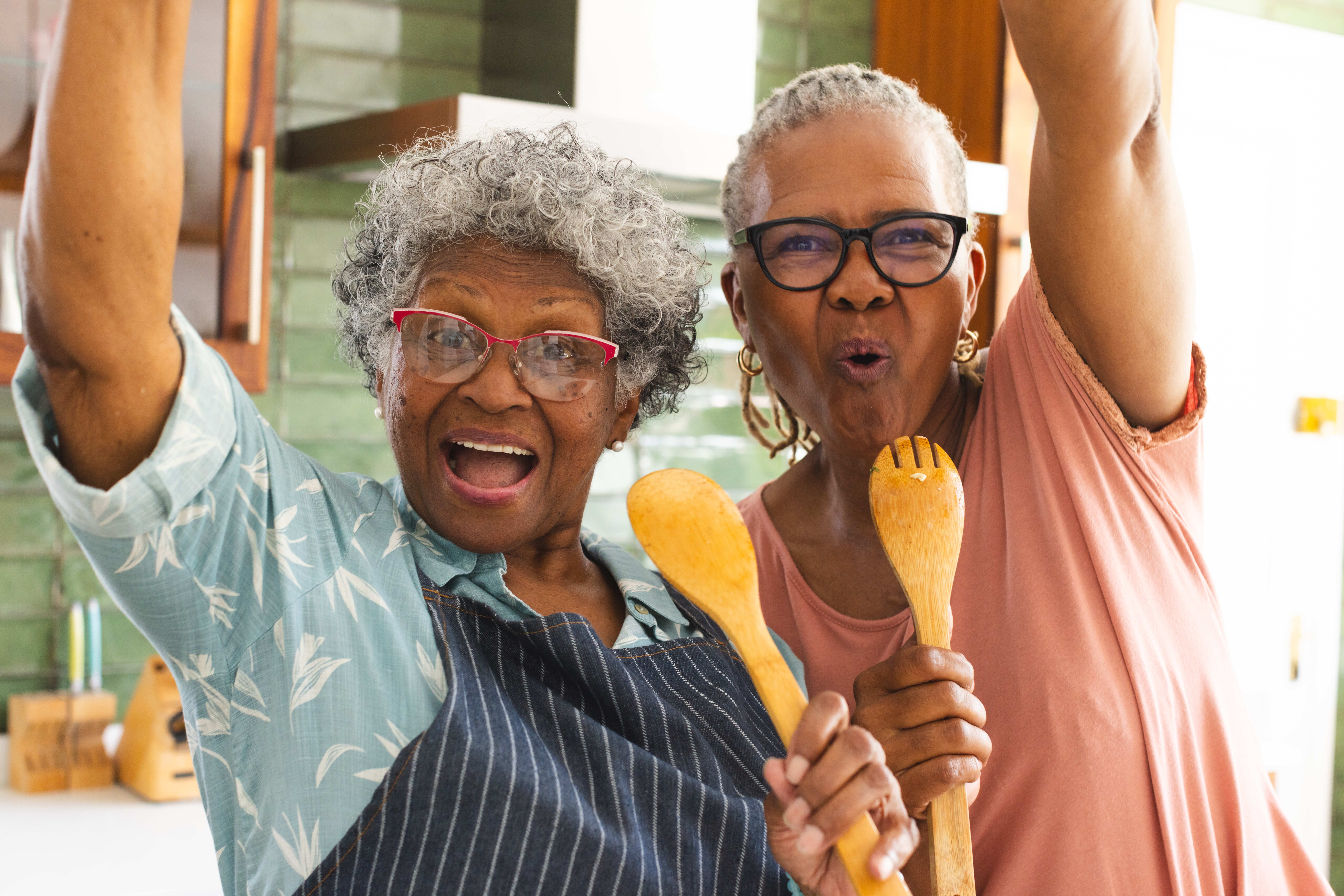 Two joyful older women in a kitchen, holding wooden utensils and smiling energetically at the camera.