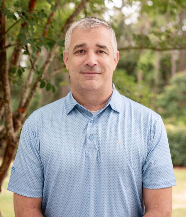 A man in a light blue polo shirt stands outdoors in front of green trees, smiling slightly at the camera.