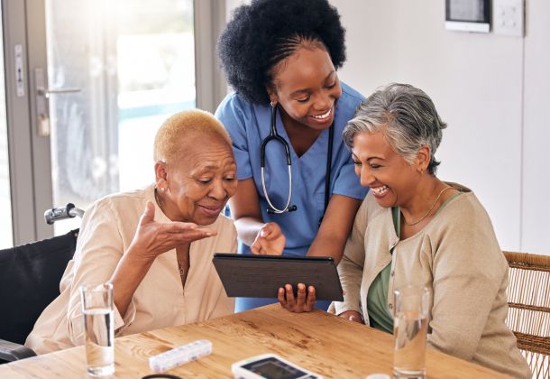 Two senior women smiling at a table with a nurse holding a tablet.
