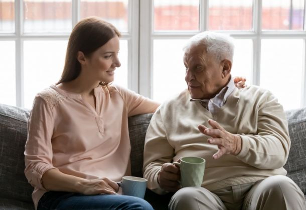 A young woman and an elderly man sitting on a sofa talking while holding mugs.