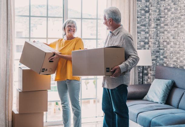 Smiling older couple holding moving boxes in a living room with stacked boxes and a large window.