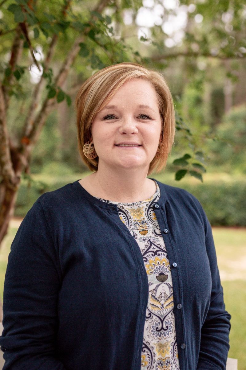 Woman with short light brown hair smiles outdoors, wearing a navy cardigan and patterned top.