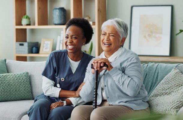 A caregiver and an older woman sitting on a sofa, both laughing.