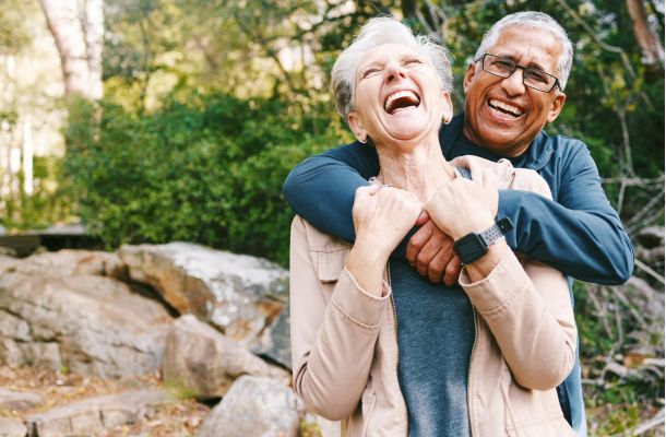 An older couple laughs joyfully as they hug each other outdoors in a wooded park.