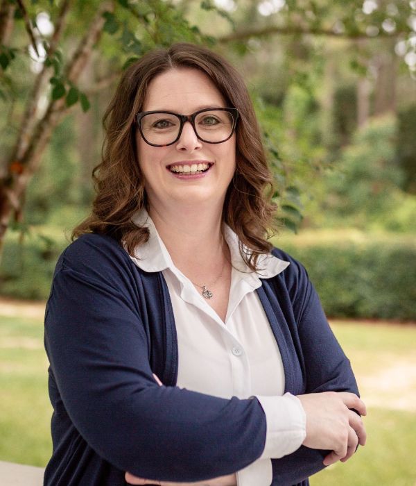 Woman with glasses smiling, arms crossed, standing outdoors with green trees and grass in the background.