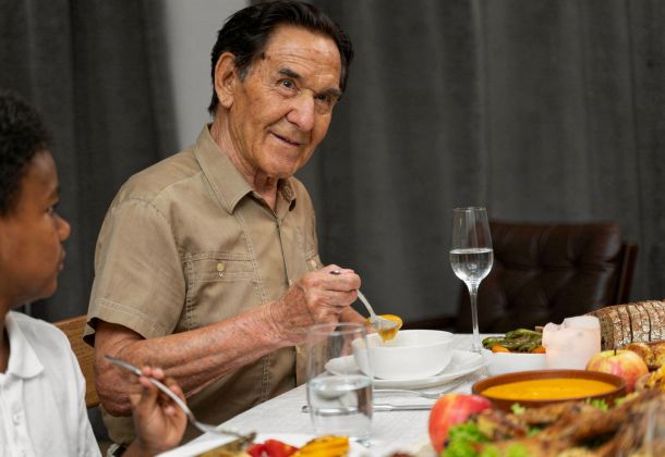 An elderly man eats soup at a dinner table, sitting beside a child, with food and drinks in front of them.