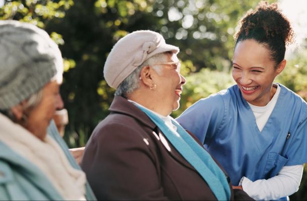 A nurse smiles and talks with two elderly women sitting outside on a bench in a garden.