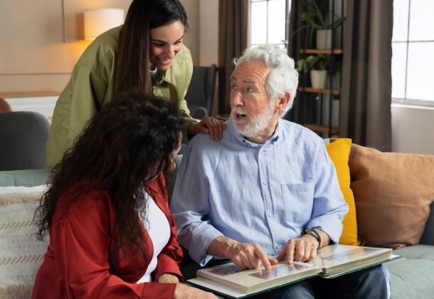 Older man showing a photo album to two women on a couch, all smiling and engaged in conversation.