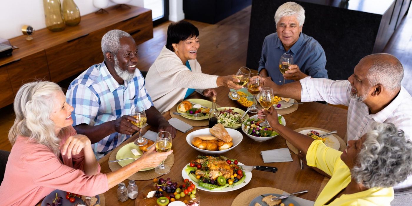 Six older adults sitting around a dining table, smiling and toasting with drinks over a meal.