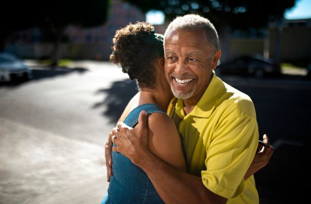 An older man in a yellow shirt smiles while hugging a woman outdoors on a sunny day.