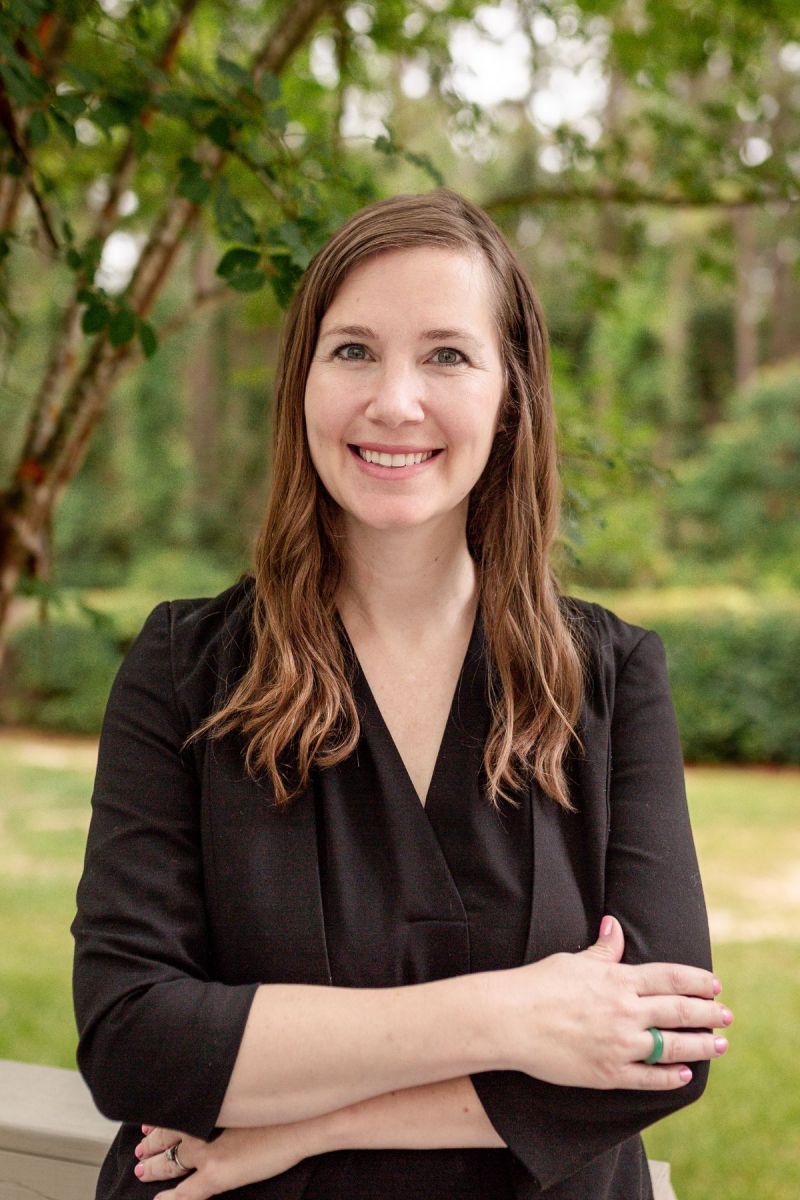 Woman with long brown hair, wearing a black top, smiling with arms crossed outdoors in a green park.