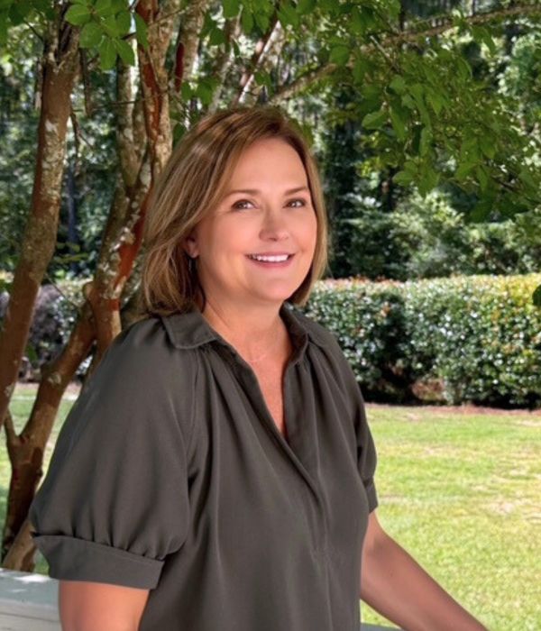 Woman with light brown hair, wearing a dark blouse, smiling outdoors near a white fence and green trees.