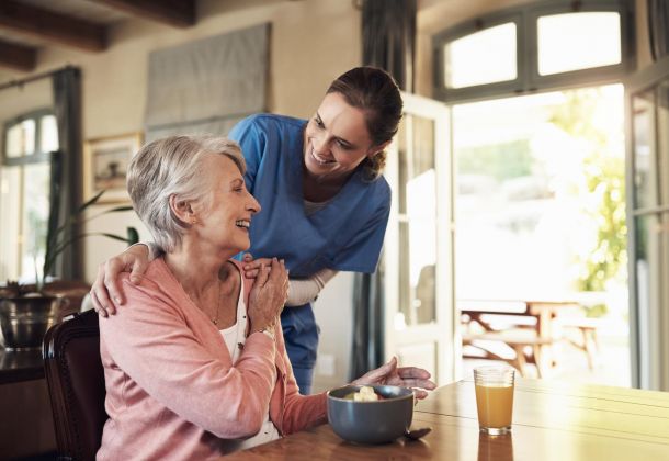 Caregiver in blue scrubs smiling at a seated elderly woman at a table with a bowl and a glass of juice.