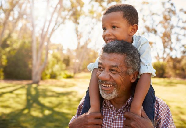 Smiling older man gives a young boy a piggyback ride outdoors on a sunny day in a park.