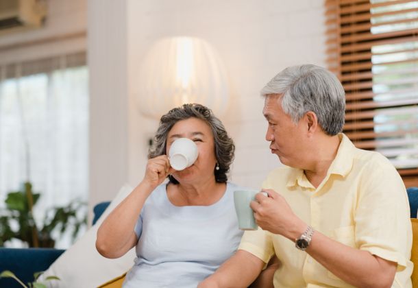 Older couple sitting on a couch at home, enjoying hot drinks and smiling together.