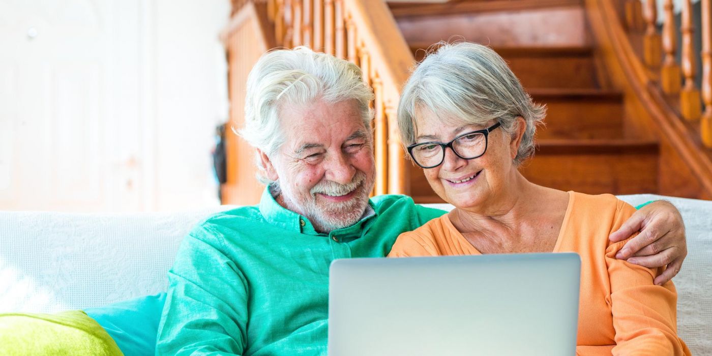 Smiling older couple sitting on a couch, looking at a laptop together in a cozy, sunlit living room with wooden stairs in the background.