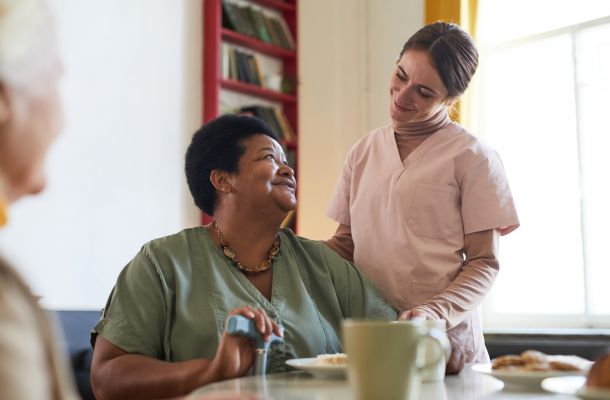 Smiling caregiver stands beside a seated woman, both sharing a warm interaction in a cozy room.