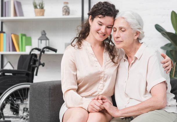 Young woman hugging elderly woman on couch, wheelchair in background, both smiling and holding hands.