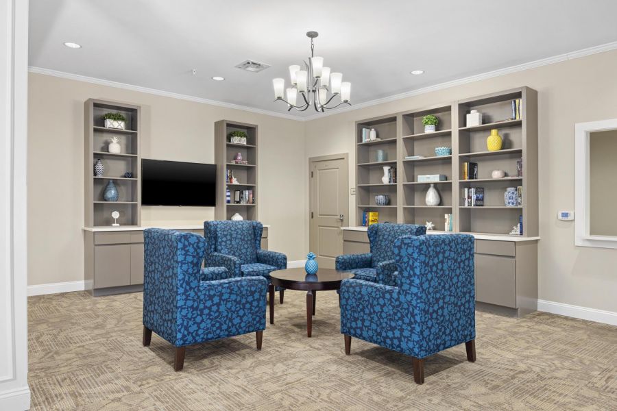 Room with blue patterned armchairs, round table, built-in shelves, and a wall-mounted TV under a chandelier.