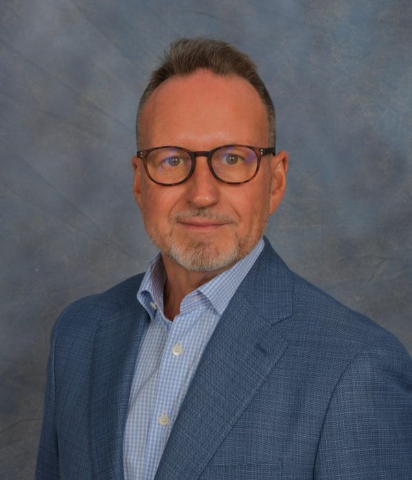 Man wearing glasses and a blue suit jacket, posing in front of a blue and gray mottled background.