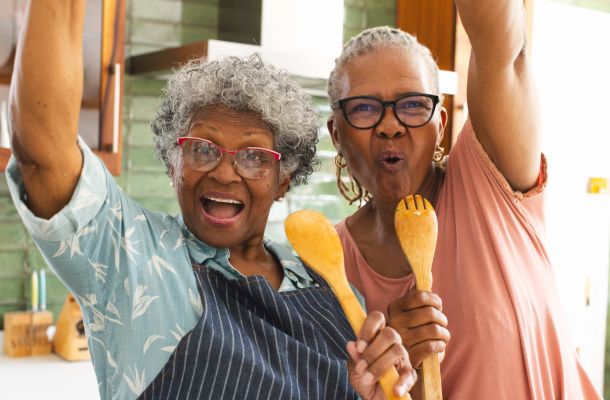 Two joyful older women in a kitchen, holding wooden utensils and smiling energetically at the camera.