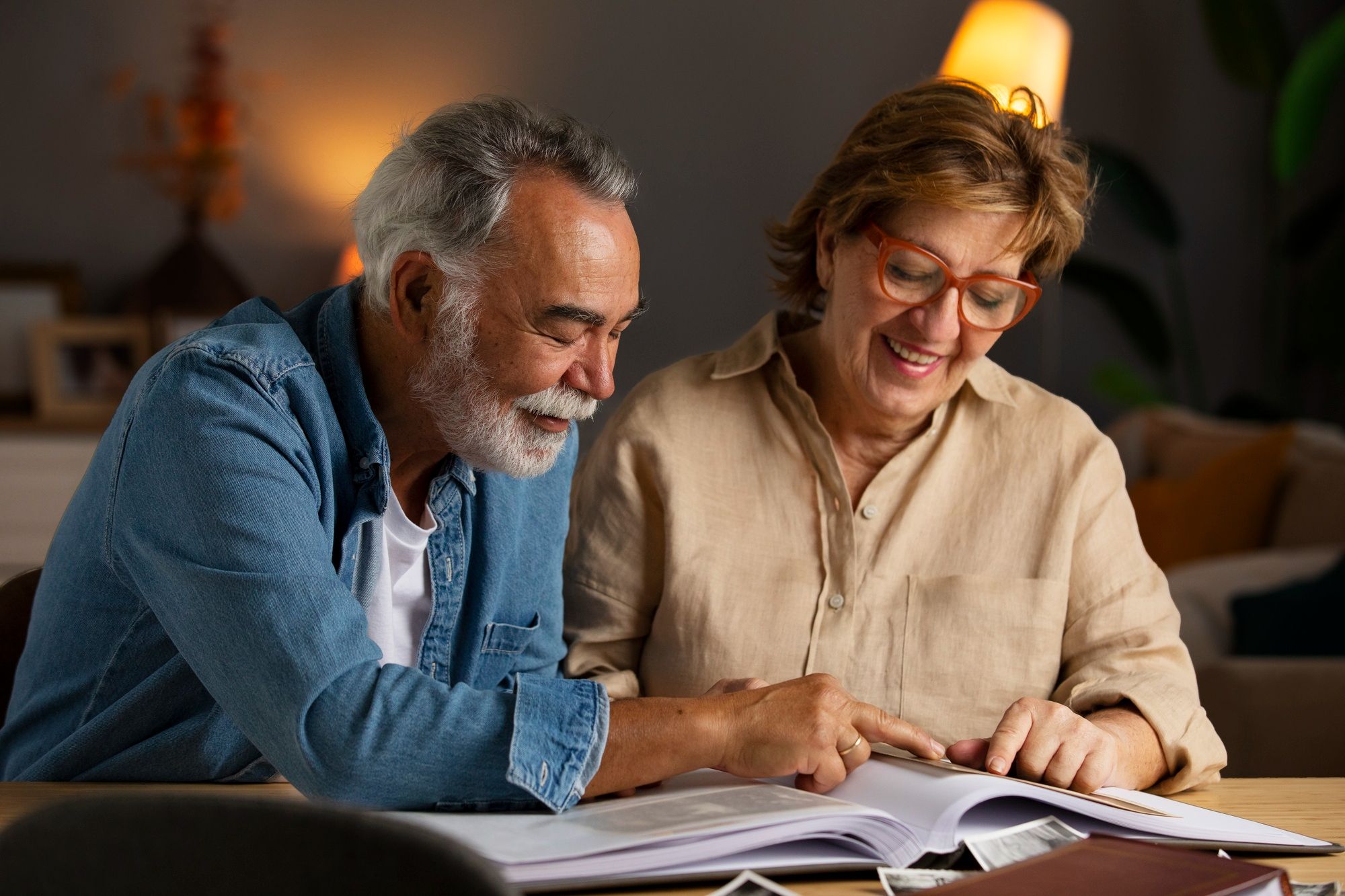 An older couple sits at a table, smiling and reading a book together in a cozy, warmly lit room.