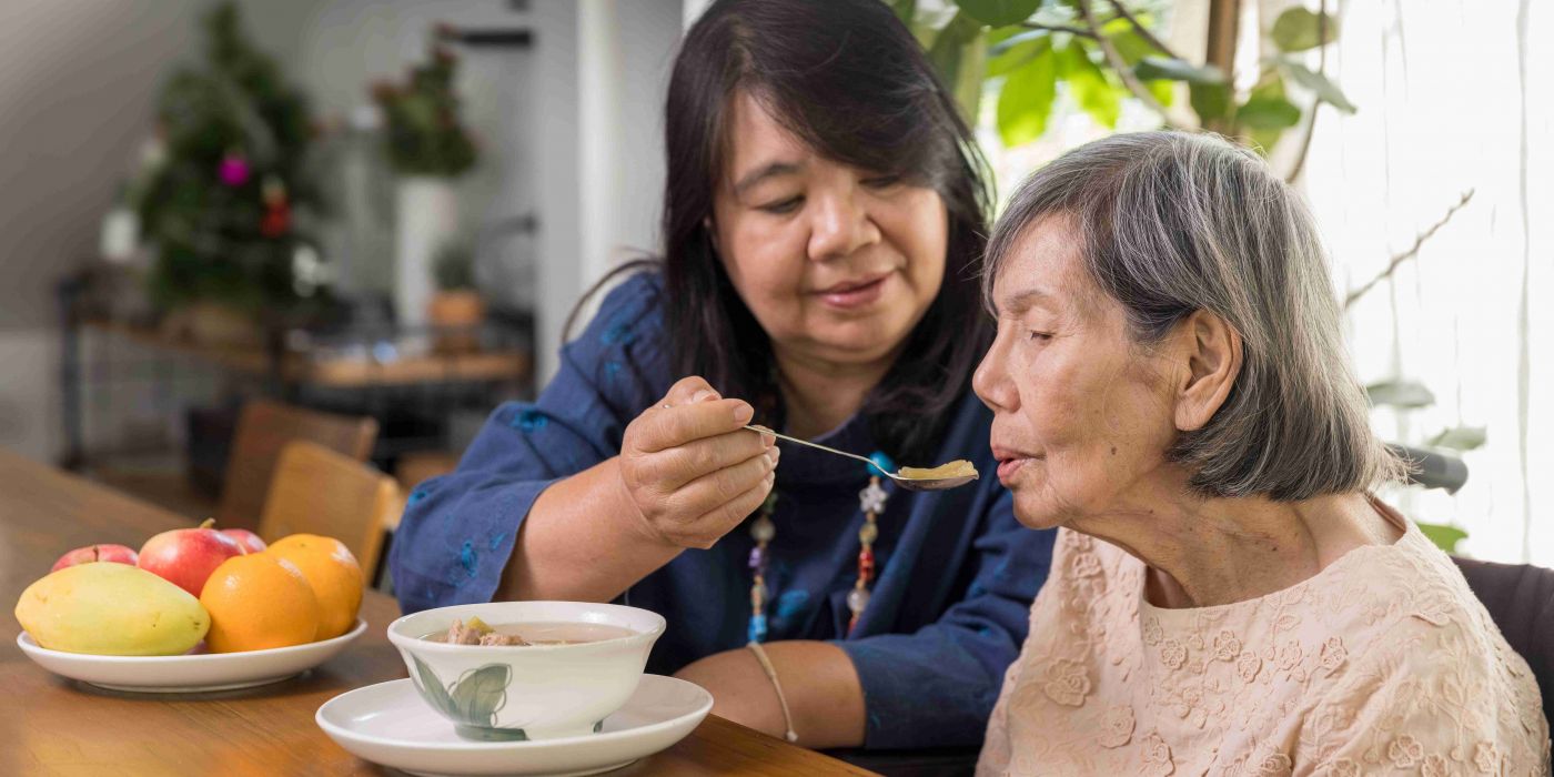 Woman feeding an elderly lady at a table with a bowl of soup and a plate of fruit.