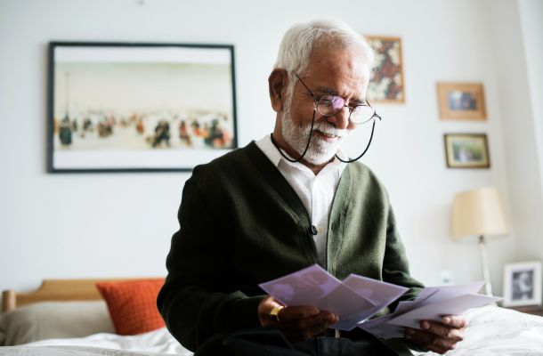 An elderly man with a white beard and glasses smiles while looking at old photographs in his bedroom.