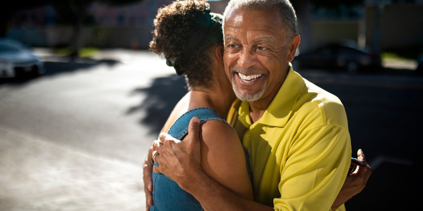 An older man in a yellow shirt smiles while hugging a woman outdoors on a sunny day.