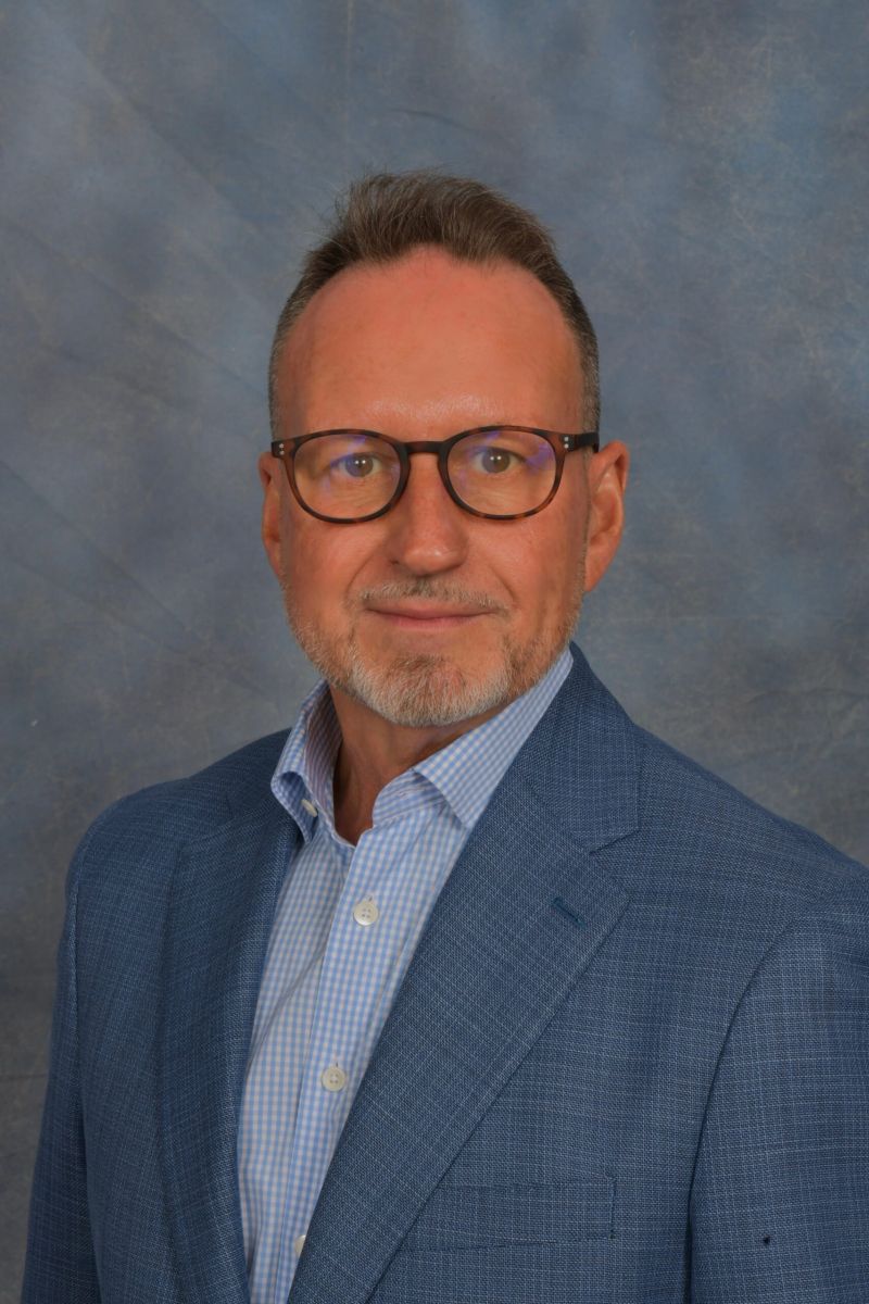 Man wearing glasses and a blue suit jacket, posing in front of a blue and gray mottled background.