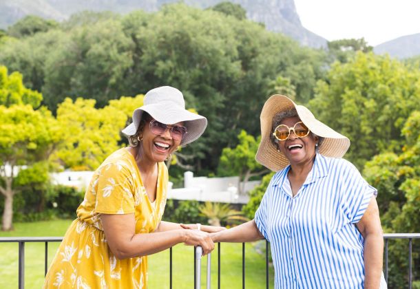 Two older women in sun hats and sunglasses smile and hold hands outdoors with trees in the background.