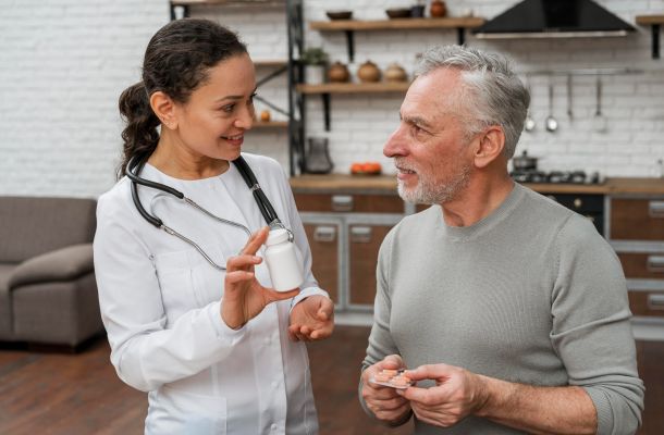 A female doctor shows a pill bottle to an older man in a kitchen, both smiling and talking.